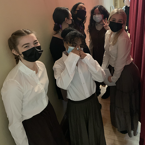 a group of masked teens in costume stand behind a curtain, smiling at the camera