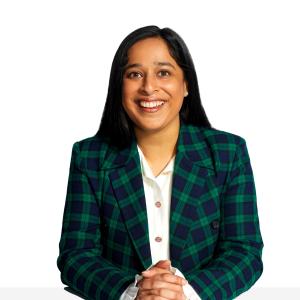 Prerna has medium toned skin, dark long hair, and she wears a white collared shirt and a green plaid blazer. She smiles at the camera and clasps her hands together.