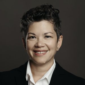 Cynthia has light skin, dark short curly hair with flecks of grey, and wears a white collared shirt and suit jacket. She smiles to the camera.