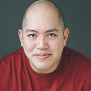 Mickey is a Filipino man, with dark eyes and is bald. He wears a red shirt and smiles into the camera.