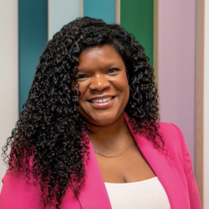 Jayan is a Black woman with long curly black hair, a white shirt, and a pink blazer. She smiles at the camera against a striped coloured background.