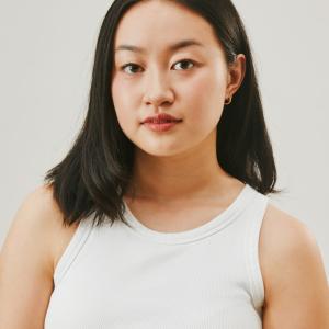 A photo of a women wearing a white tank top, with short black hair looks at the camera.
