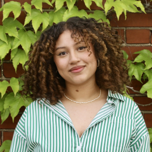 A person with curly hair stands in front of a brick wall smiling at the camera. 