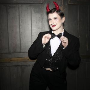 A person wearing red devil and a 3 piece sparkly suit ears grins cheekily at the camera while holding onto their bowtie
