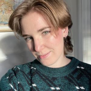 A person wearing a green and black printed sweater sits in a sunny spot smiling at the camera