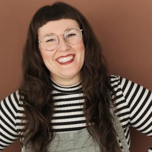 A person wearing a striped shirt overalls stands with hands on hip smiles at the camera