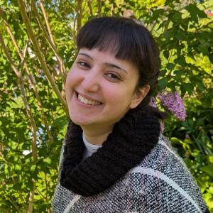A women standing in nature wears a black turtle neck and smiles happily at the camera 