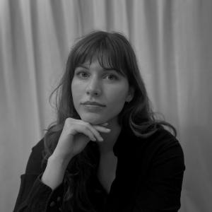 A women in a black long sleeve sits with her hand under her chin as she looks at the camera stoically 
