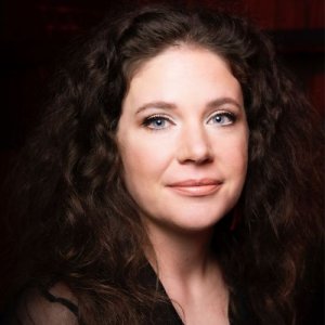 Damien Nelson's headshot, a woman with pale skin and dark curly hair against a dark background