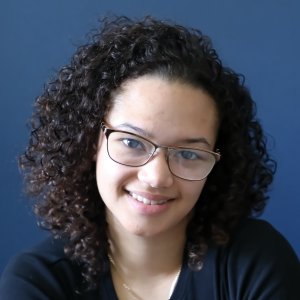 Kayleigh has dark curly hair, glasses, and is smiling at the camera against a blue background