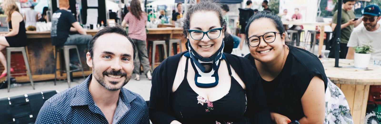 Three friends gather at the Fringe patio, all smiling at the camera, one sits in a wheelchair and wears a neckbrace