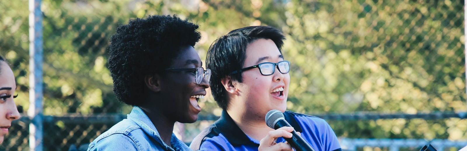 Two teens animatedly speak into a microphone, with trees and leaves in the background