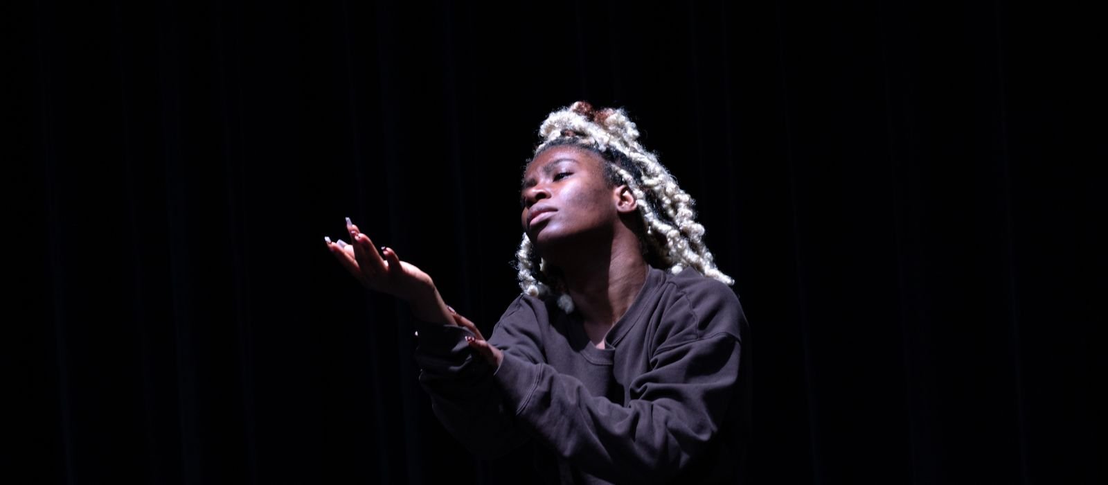 A Black woman with blond dreads holds her hands up and looks up, against a black background