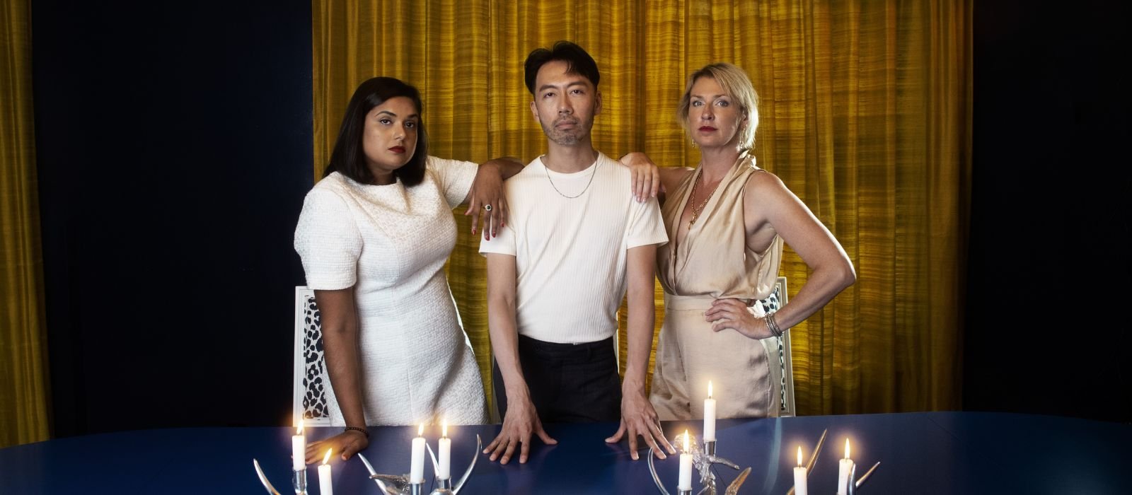 Three comedians stand with their arms around each other, with a gold curtain in the background and table with lit candles in the foreground.