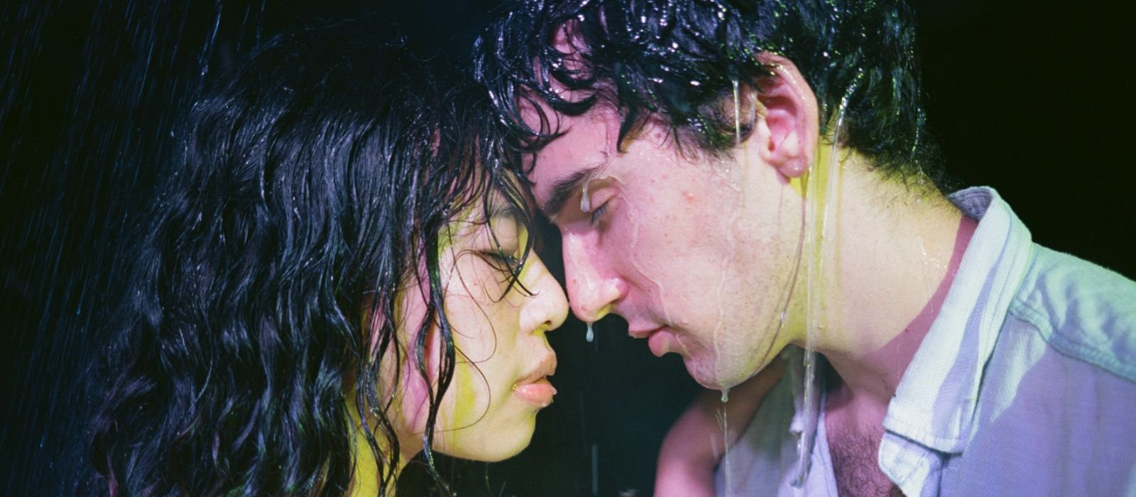 A woman and a man face each other with their noses touching, water running down their hair and faces.
