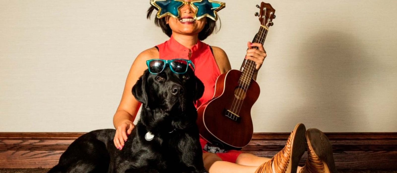 Photograph of Vivian Chong sitting on the floor smiling, wearing big star-shaped sunglasses and holding a ukulele. Beside her is guide dog Catcher, lying regally at her side, with star-shaped sunglasses on his head.