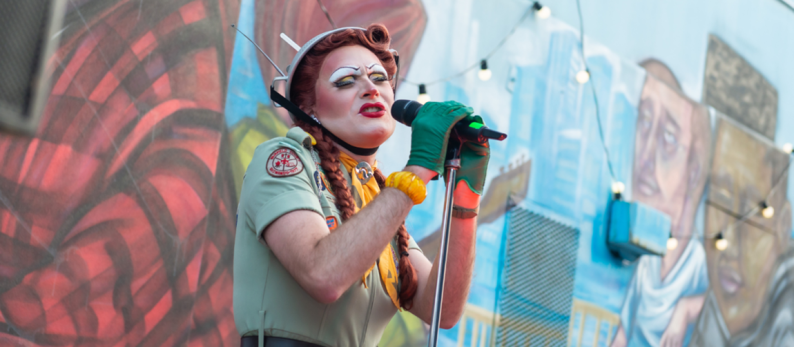 Pearle Harbour, a drag performer, has a metal colander on her head and sings to the audience from an outdoor stage