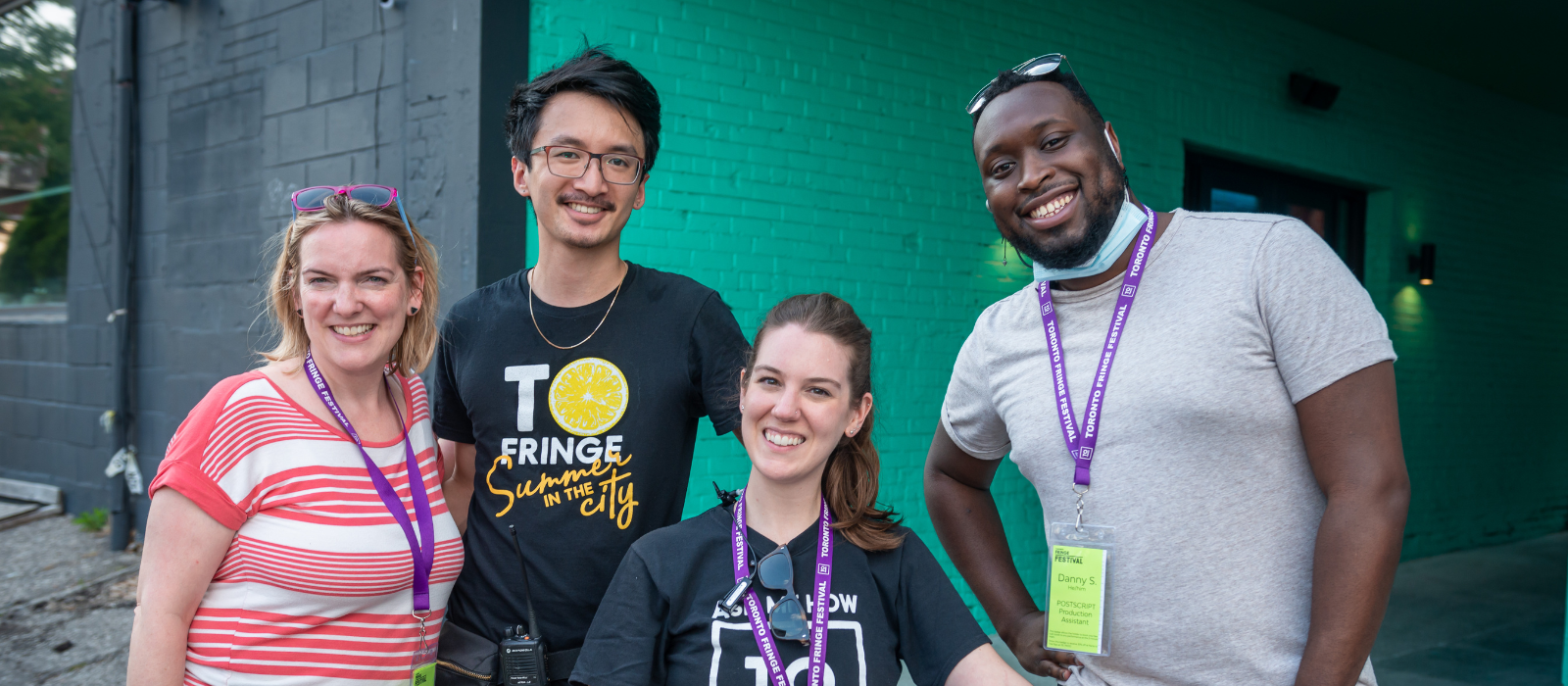 Four Fringe staff members stand together at the Patio, smiling at the camera, with their staff shirts and lanyards visible