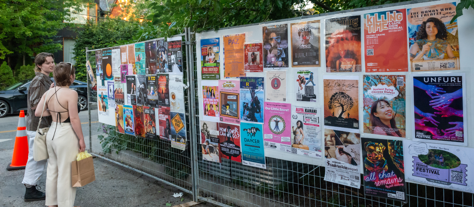 Two patrons look at a wall of Fringe show posters at the 2023 Fringe patio