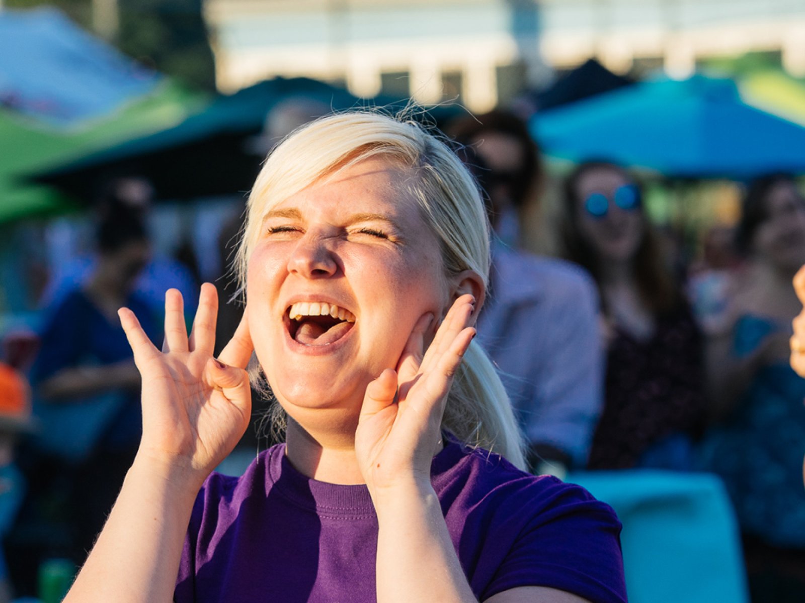 A female fringe volunteer holds her hands to her mouth as she yells with enthusiasm at the Fringe patio